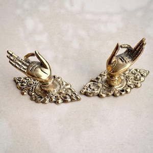 May include: A pair of antique brass hand-shaped cabinet pulls. Each pull depicts a detailed hand gesture atop an ornate, decorative base. The brass has a warm, aged patina, enhancing the vintage look. Perfect for adding a unique touch to furniture.