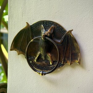 May include: A bronze-colored bat-shaped door knocker. The bat has outstretched wings and a ring for knocking. The knocker is mounted on a light-colored wall. The bat's wings have a decorative pattern.