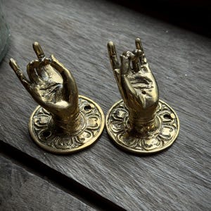 May include: Two brass-coloured decorative hand-shaped door pulls. Each hand is in a rock-on gesture, mounted on a round base with an ornate floral design. The hardware is set against a weathered wooden surface.
