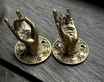May include: Two brass wall hooks shaped like hands making a gesture with the index and pinky fingers extended. Each hand is mounted on a circular base featuring a decorative floral relief pattern and two small holes for mounting screws.