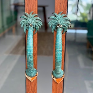 May include: A pair of vertical door handles shaped like palm trees, featuring a weathered teal and green patina finish. Each handle is mounted onto a vertical wooden door frame with a distinct grain pattern and a metallic base plate at the bottom.