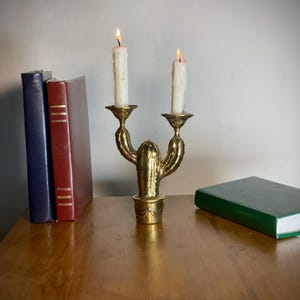 May include: A brass cactus-shaped candle holder with two lit white candles. The candle holder sits on a wooden surface next to two books and a green book. The candles are burning with a warm glow.