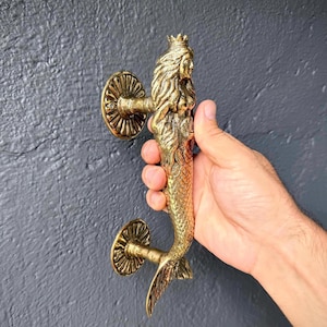 May include: A brass mermaid door handle. The handle features a mermaid with a crown, flowing hair, and a scaled tail. The handle is held by a hand, and the door hardware is mounted on a dark gray surface.