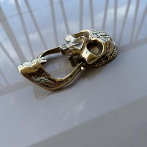 May include: A brass-colored belt buckle in the shape of a skull. The skull has detailed features, including eye sockets, a nose cavity, and teeth. The buckle has a curved bar for the belt to pass through.