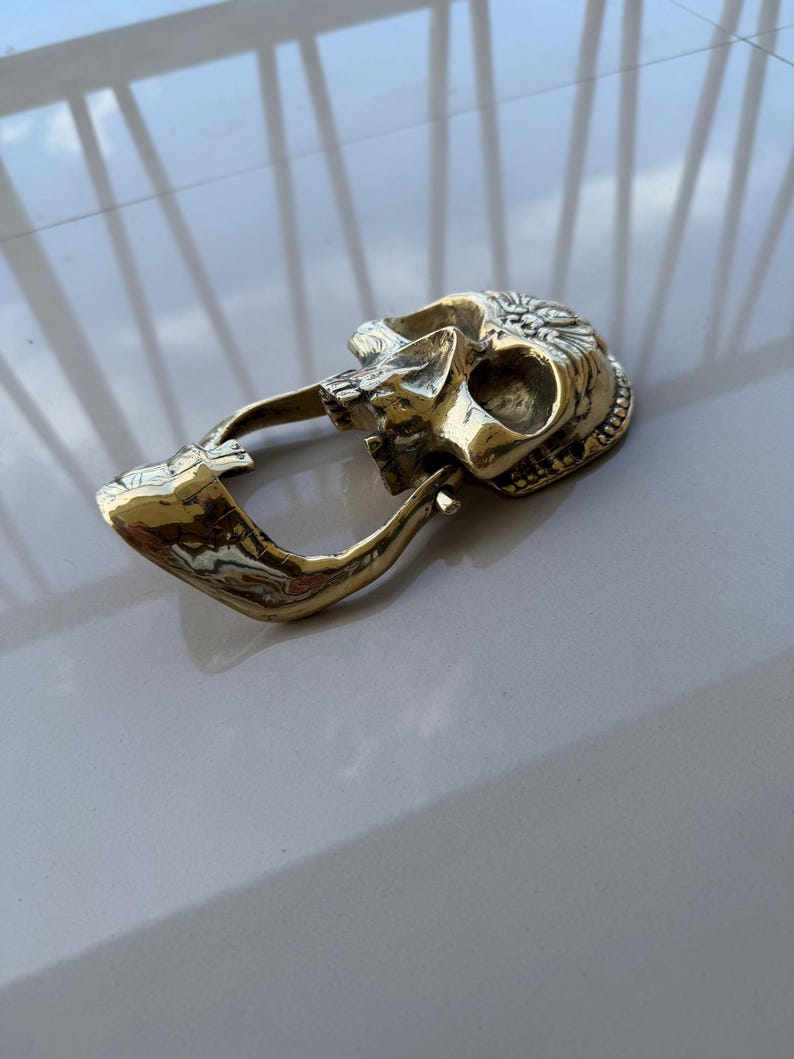 May include: A brass-colored belt buckle in the shape of a skull. The skull has detailed features, including eye sockets, a nose cavity, and teeth. The buckle has a curved bar for the belt to pass through.