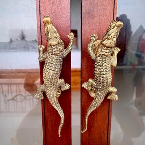 May include: A pair of brass-colored metal door handles shaped like crocodiles, mounted vertically on two separate dark wood door panels. Each handle features a textured, scaled surface and a long tail extending downward along the wood.