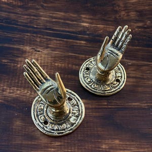 May include: Two antique brass-colored hand-shaped door pulls. Each hand is in a gesture of offering, mounted on a round, ornate base with screw holes. The background is a dark wood surface.