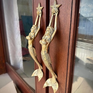 May include: A pair of polished brass door handles shaped like mermaids, each depicted with arms raised above their heads to hold a five-pointed starfish. The figures feature detailed scales on their tails and are mounted vertically on a wooden door.