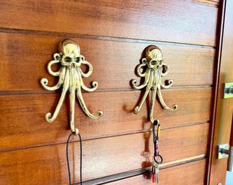 May include: Two polished brass wall hooks shaped like octopuses with human skull heads. Each hook features multiple curved tentacles extending outward to serve as hanging points. One hook holds a black cord, and the other holds a keychain.
