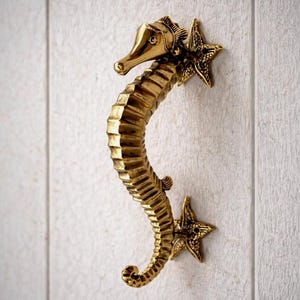 May include: A brass seahorse-shaped door handle. The handle is a golden color and is mounted on a white wooden surface. The seahorse has a detailed design, including a starfish at each end.