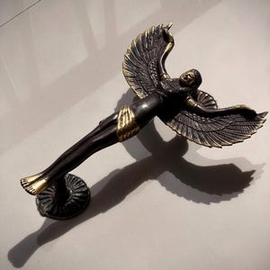 May include: A bronze-toned sculpture of a winged figure in a reclining pose. The figure has outstretched arms and detailed wings with gold accents. The sculpture is mounted on a circular base and casts a long shadow.