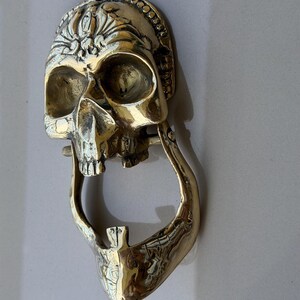 May include: A brass skull-shaped door knocker with intricate details. The knocker features a detailed skull design with an open mouth and a decorative crown. The knocker is mounted on a light-colored surface.