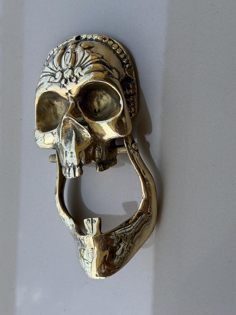 May include: A brass skull-shaped door knocker with intricate details. The knocker features a detailed skull design with an open mouth and a decorative crown. The knocker is mounted on a light-colored surface.