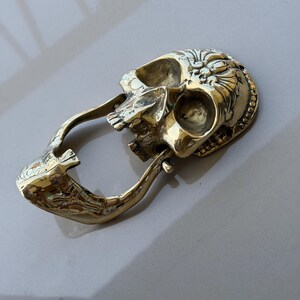 May include: A brass-colored skull-shaped keychain with an open mouth and a loop for attaching keys. The skull has detailed carvings on the forehead and around the eye sockets. The keychain is made of a shiny, metallic material.