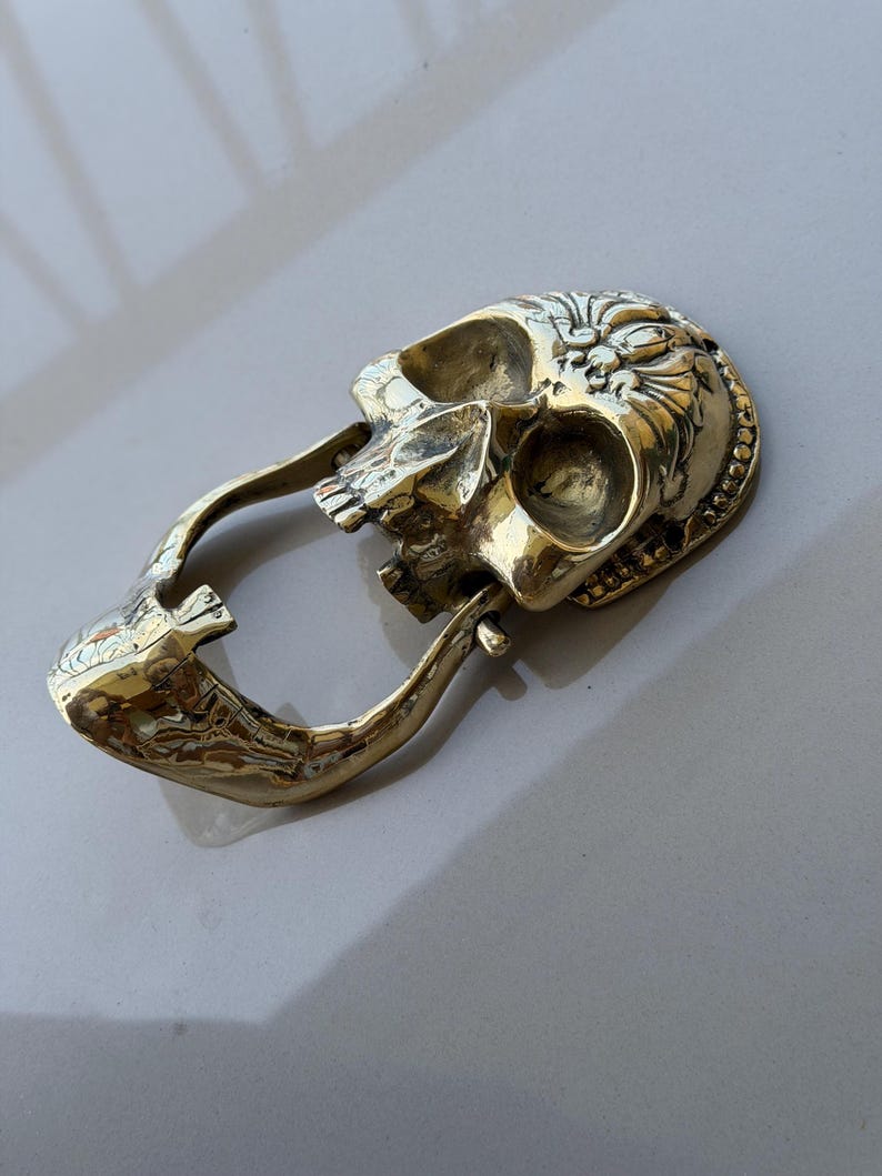 May include: A brass-colored skull-shaped keychain with an open mouth and a loop for attaching keys. The skull has detailed carvings on the forehead and around the eye sockets. The keychain is made of a shiny, metallic material.
