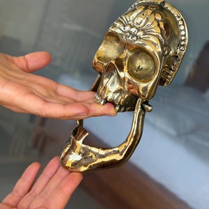 May include: A brass skull door knocker with intricate floral detailing on the forehead. The knocker is a polished gold color and features a hinged jaw. The skull's eye sockets and nasal cavity are visible. The knocker is designed to be mounted on a door.