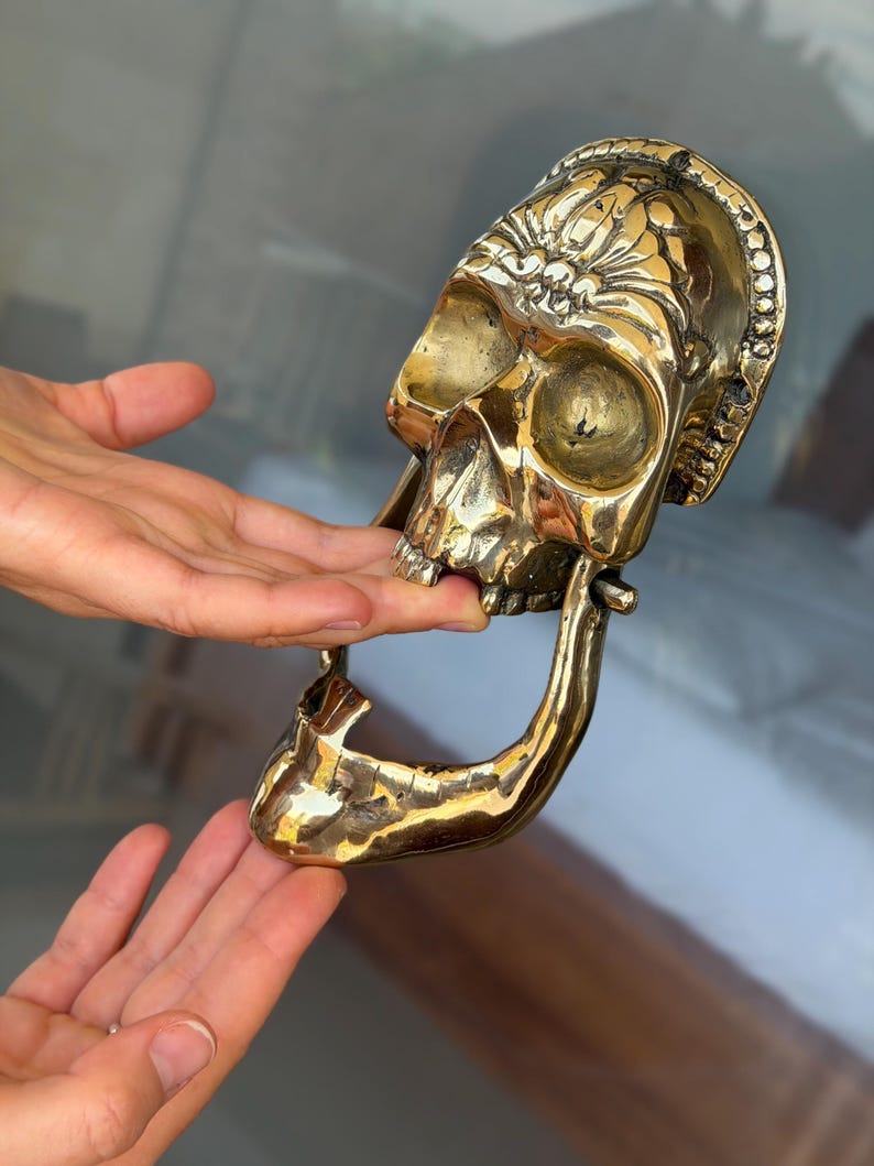 May include: A brass skull door knocker with intricate floral detailing on the forehead. The knocker is a polished gold color and features a hinged jaw. The skull's eye sockets and nasal cavity are visible. The knocker is designed to be mounted on a door.