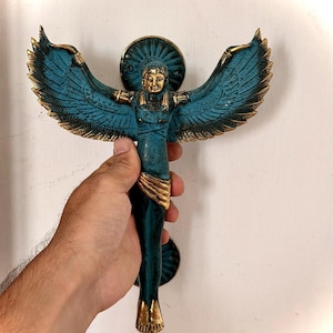 May include: A metal door handle shaped like a winged figure with a teal and gold finish. The figure has outstretched wings, a detailed face, and a draped garment around the waist. The handle features circular mounting plates at the top and bottom.