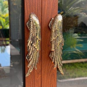 May include: A pair of vertical door handles crafted from polished brass, each shaped like a detailed bird wing with textured feathers. The handles are mounted onto a dark brown wood-grain door frame, featuring a reflective metallic finish.