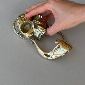 May include: A brass skull-shaped door knocker with intricate details. The knocker features a skull design with a hinged handle. The brass has a polished finish, reflecting light. The skull is held by a hand.