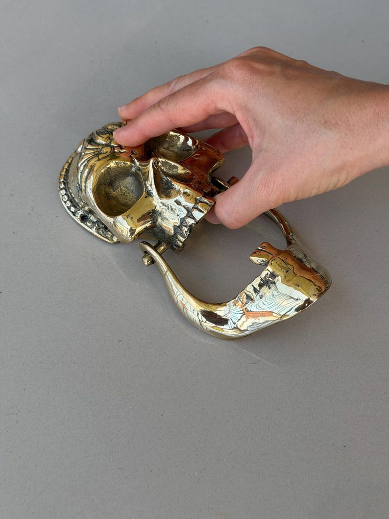 May include: A brass skull-shaped door knocker with intricate details. The knocker features a skull design with a hinged handle. The brass has a polished finish, reflecting light. The skull is held by a hand.
