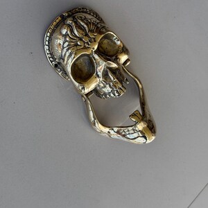 May include: A brass skull-shaped door knocker with intricate detailing. The knocker features a detailed skull design with a decorative border and a curved handle. The metal has a polished, golden finish, adding a touch of gothic style.