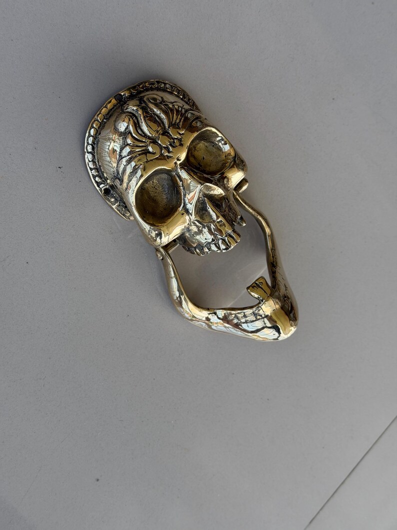May include: A brass skull-shaped door knocker with intricate detailing. The knocker features a detailed skull design with a decorative border and a curved handle. The metal has a polished, golden finish, adding a touch of gothic style.