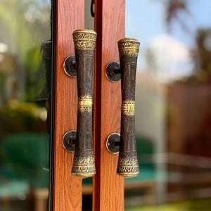 May include: A pair of vertical door handles mounted on a wood-grain door. Each handle features a dark, cylindrical center with intricate, engraved patterns and polished gold-tone metal accents at the top, middle, and bottom sections.