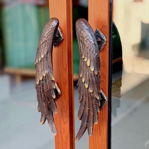 May include: Two door handles shaped like feathered wings, finished in a dark bronze tone with gold highlights. The handles are mounted vertically on a wooden door frame with a visible wood grain pattern, featuring detailed sculpted feather textures.