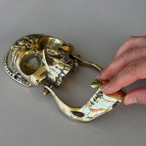 May include: A brass skull-shaped belt buckle with intricate detailing. The buckle features a skull design with an open mouth and a curved, hook-like closure. The metal has a polished, golden finish.