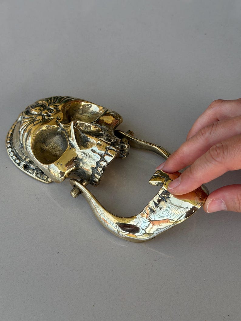 May include: A brass skull-shaped belt buckle with intricate detailing. The buckle features a skull design with an open mouth and a curved, hook-like closure. The metal has a polished, golden finish.