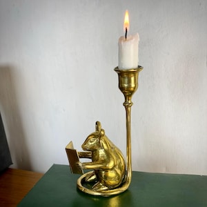 May include: A brass candle holder featuring a detailed sculpture of a squirrel reading a book. A lit white candle sits atop the holder. The candle holder is on a green surface.