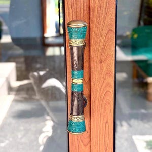May include: A vertical door handle mounted on a wood-grain door. The handle features a cylindrical design with alternating sections of dark brown metal and bright turquoise metal, accented by thin gold-colored bands with intricate engraved patterns.