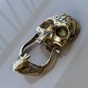 May include: A detailed, golden skull-shaped door knocker. The knocker features intricate carvings and a hinged jaw, creating a unique and decorative element for any door. The skull is made of a shiny, metallic material.