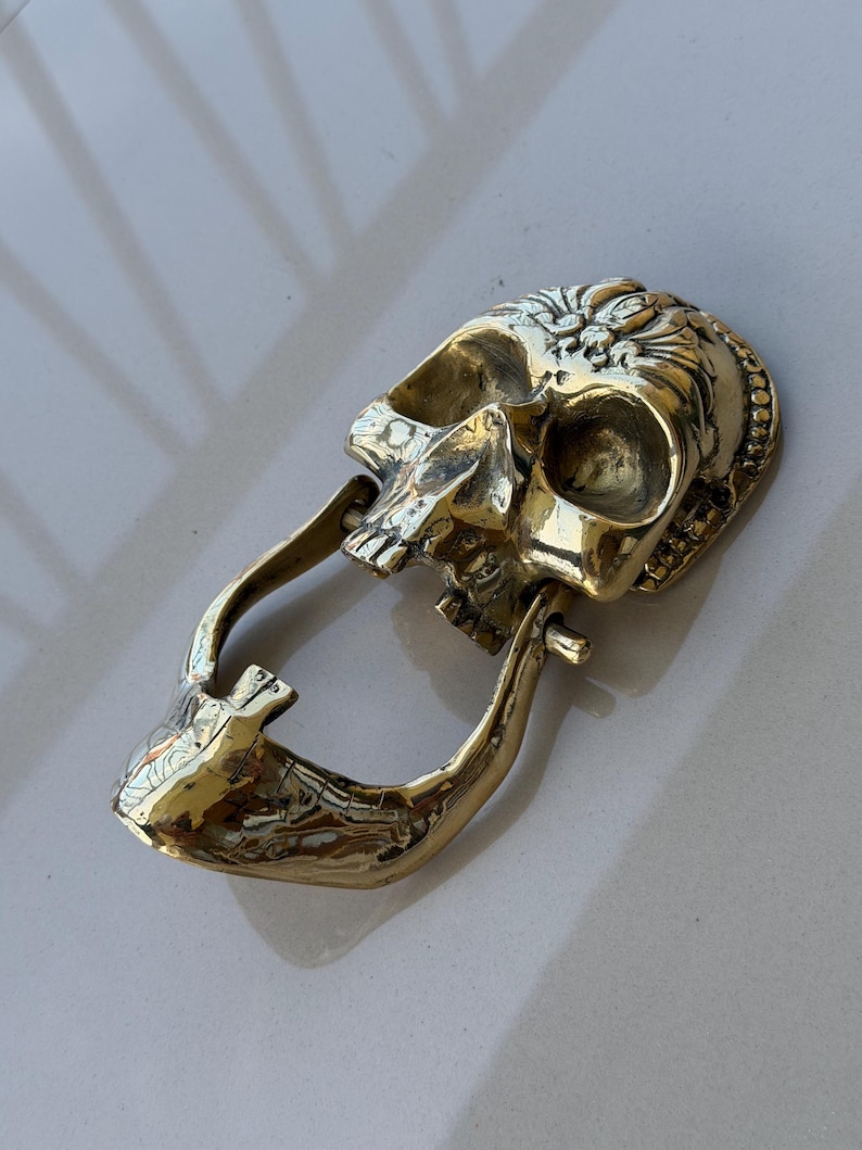 May include: A detailed, golden skull-shaped door knocker. The knocker features intricate carvings and a hinged jaw, creating a unique and decorative element for any door. The skull is made of a shiny, metallic material.