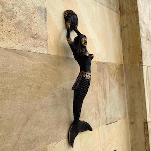 May include: A bronze-colored mermaid sculpture with a seashell headdress, arms raised. The mermaid has a scaled tail and is mounted on a textured wall. The sculpture is a decorative accent piece.