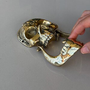 May include: A brass skull-shaped pipe with intricate details. The pipe features a skull design with a detailed face and a curved pipe stem. The brass has a polished, golden finish, and the pipe is resting on a light gray surface.