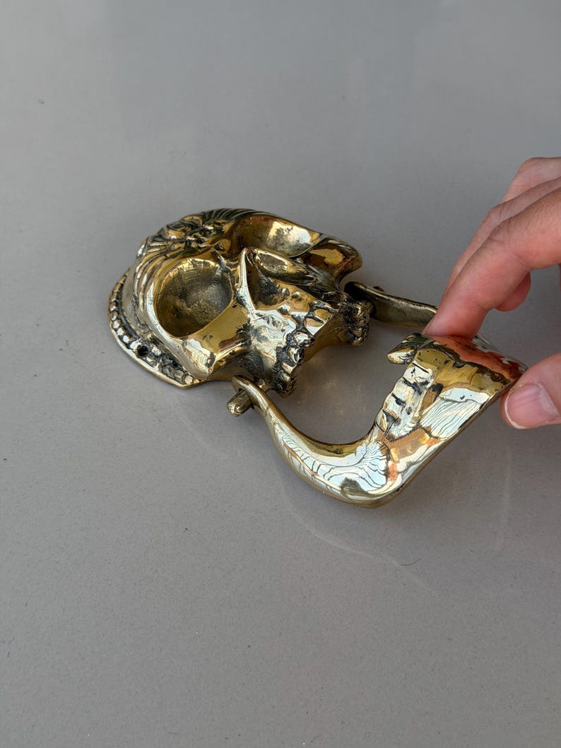 May include: A brass skull-shaped pipe with intricate details. The pipe features a skull design with a detailed face and a curved pipe stem. The brass has a polished, golden finish, and the pipe is resting on a light gray surface.
