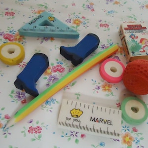 Può includere: Una collezione di gomme da cancellare vintage con forma di stivale blu, palla rossa, anello verde e bianco, anello rosa e bianco, righello triangolare blu con la parola "MARVEL" stampata, righello blu e bianco con le parole "MARVEL" e "for you" stampate, e una matita colorata. Le gomme da cancellare sono su uno sfondo floreale.