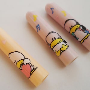 Może przedstawiać: Trzy różowe i żółte plastikowe nakładki na długopisy z motywem kreskówkowej kaczki. Każda nakładka przedstawia inną kaczkę z innym wyrażeniem twarzy i dodatkiem. Kaczki noszą różowe serce, niebieską nutę muzyczną i żółtą gwiazdę.