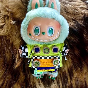 May include: A plush doll with a mint green bunny hood and a lime green jacket. The doll has a smiling face with large eyes and a set of teeth. The jacket has decorative jewels and the text "MOCK-UP CONTROL". The doll's arms have black and white checkered sleeves.