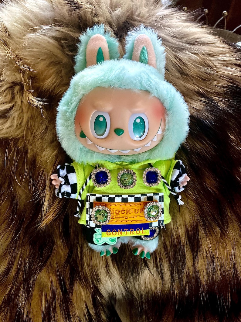 May include: A plush doll with a mint green bunny hood and a lime green jacket. The doll has a smiling face with large eyes and a set of teeth. The jacket has decorative jewels and the text "MOCK-UP CONTROL". The doll's arms have black and white checkered sleeves.