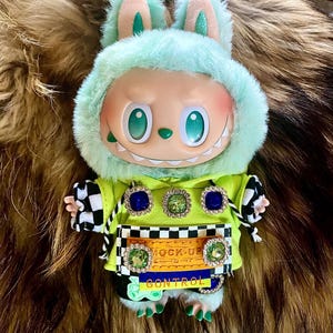 May include: A plush doll with a mint green hooded jacket and bunny ears. The doll has a smiling face with large green eyes and a row of teeth. The jacket is adorned with jewel-like embellishments and the text "MOCK-UP CONTROL".