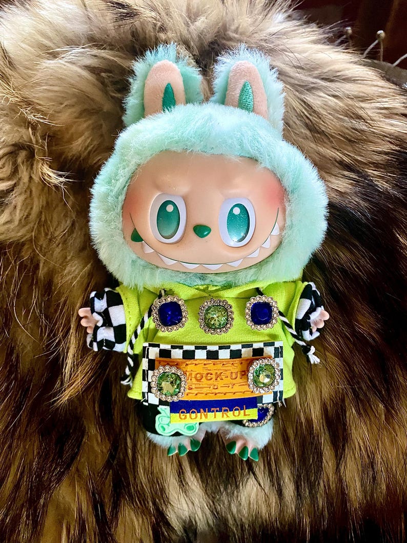 May include: A plush doll with a mint green hooded jacket and bunny ears. The doll has a smiling face with large green eyes and a row of teeth. The jacket is adorned with jewel-like embellishments and the text "MOCK-UP CONTROL".