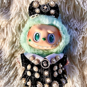 May include: A whimsical doll with a mint green furry hood and a black dress adorned with pearls and rhinestones. The doll has large, expressive eyes and a decorative bow on its head, creating a unique and fashionable aesthetic.
