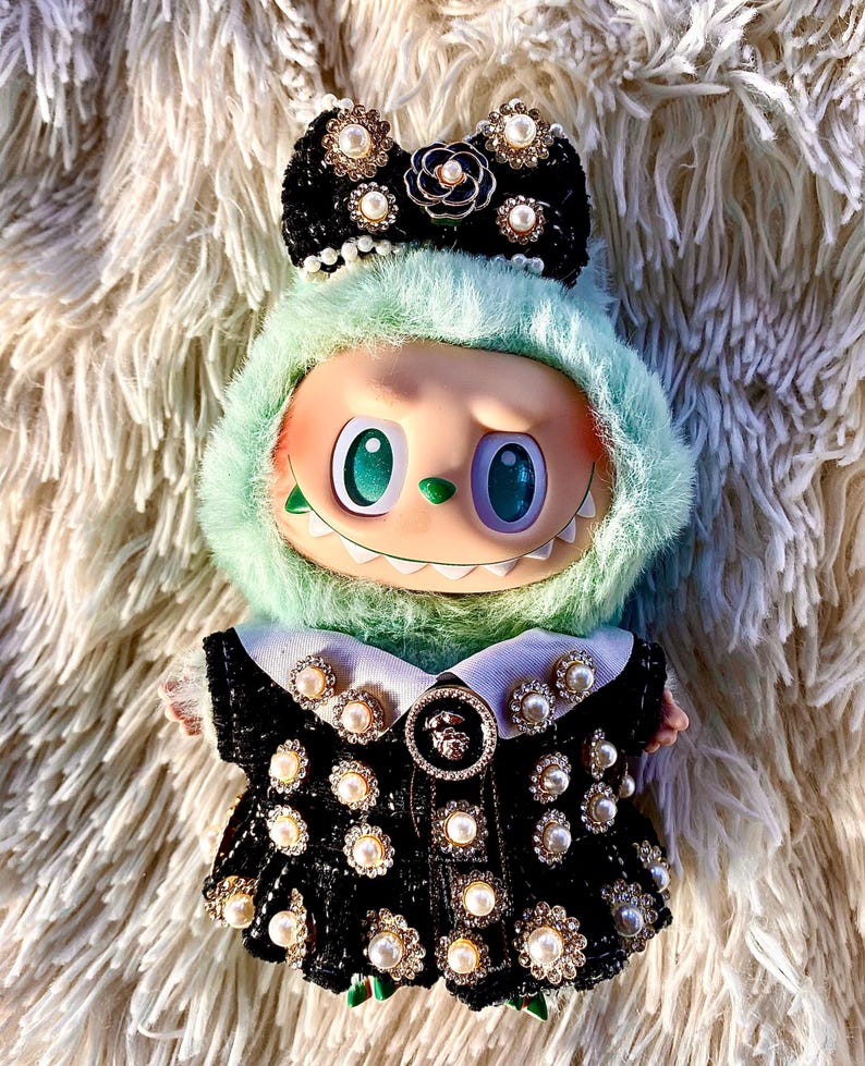May include: A whimsical doll with a mint green furry hood and a black dress adorned with pearls and rhinestones. The doll has large, expressive eyes and a decorative bow on its head, creating a unique and fashionable aesthetic.