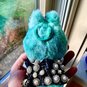 May include: A teal-colored, fluffy keychain with a black and white patterned dress adorned with faux pearls and rhinestones. The keychain has a green clasp and a small tag that reads "POP".