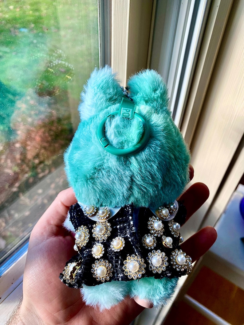 May include: A teal-colored, fluffy keychain with a black and white patterned dress adorned with faux pearls and rhinestones. The keychain has a green clasp and a small tag that reads "POP".