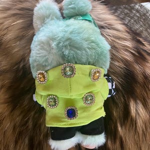 May include: A plush bunny toy with a mint green fur body and a matching pom-pom hat. The bunny wears a lime green jacket adorned with jewel-like buttons. Black and white checkered fabric accents the toy's design.