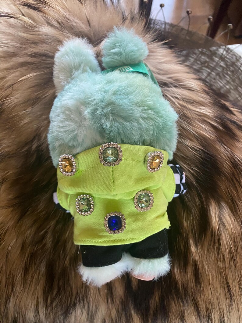May include: A plush bunny toy with a mint green fur body and a matching pom-pom hat. The bunny wears a lime green jacket adorned with jewel-like buttons. Black and white checkered fabric accents the toy's design.